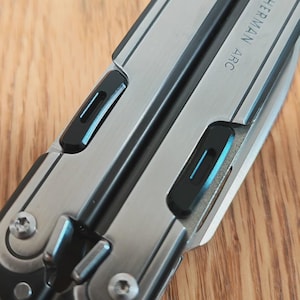 Puede incluir: Primer plano de una multiherramienta con un diseño plateado y negro. La herramienta tiene el texto "LEATHERMAN ARC" impreso en el lateral. La herramienta está sobre una superficie de madera.