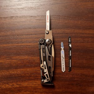 Op de afbeelding: Een zilver en zwart multi-tool met een zaagblad uitgeschoven, naast twee extra zaagbladen. De bladen zijn zilver en zwart, met tekst. Op een houten oppervlak.
