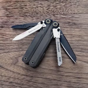 May include: A black multi-tool with several open blades and tools, including a saw, knife, and scissors. The multi-tool is open on a wooden surface, showcasing its various functions. The saw blade is labeled "Metal".