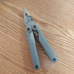 May include: A grey multi-tool featuring pliers, a knife, and various other implements. The tool incorporates orange detailing and a silver hinge. It is shown open, with the pliers visible at the top and the handle at the bottom. The tool rests on a wooden surface.