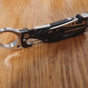 May include: A Leatherman multi-tool, with a silver ring, black handle, and silver accents. The tool is designed with multiple functions and is shown on a wooden surface. The Leatherman brand name is visible.