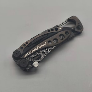 Może przedstawiać: Wielofunkcyjne narzędzie Leatherman Skeletool w kolorze ciemnoszarym. Posiada karabińczyk i różne narzędzia. Widoczna jest nazwa marki. Rękojeść ma unikalny wzór.