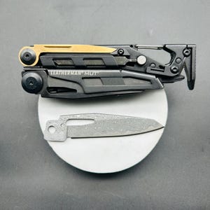 May include: A black and gold Leatherman MUT multi-tool with a silver blade. The multi-tool has a carabiner clip and a black handle. The blade is silver with a unique pattern. The text "LEATHERMAN MUT" is visible on the tool.