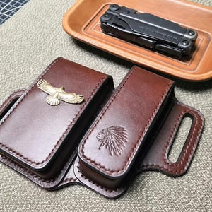 May include: A dark brown leather multi-tool sheath with two compartments, featuring a gold-toned eagle emblem and a stamped Native American headdress design. A black multi-tool rests in a separate tan leather tray above the sheath.