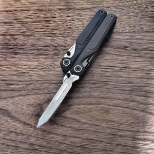 Op de afbeelding: Een zwart en grijs multitool met een uitgeschoven zilveren mes. De tool heeft een getextureerde handgreep en goudkleurige accenten. Het mes is open en toont de scherpe rand. De tool ligt op een houten oppervlak.