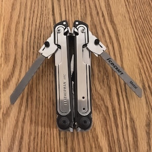 May include: A multi-tool with a silver and black finish, featuring a saw blade extended on each side. The tool is labeled "LEATHERMAN ARC" and "JCD CRAFT Metal". The tool is open, displaying its various components.