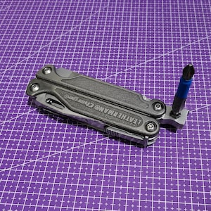 Puede incluir: Una herramienta multifunción Leatherman Charge en posición cerrada con un mango texturizado gris oscuro y detalles en metal plateado. Una punta de destornillador negra con una banda azul está insertada en el soporte, sobresaliendo lateralmente.