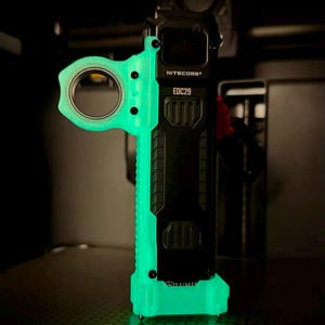 Può includere: Una torcia Nitecore EDC29 nera e verde neon. La torcia ha un'impugnatura strutturata e un'apertura circolare sul lato. Il testo "NITECORE" e "EDC29" è visibile sul dispositivo.