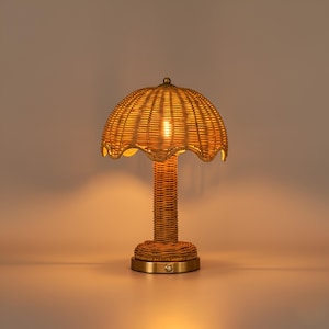 Puede incluir: Una lámpara de mesa de ratán tejido con una pantalla festoneada. La lámpara tiene una base y un tallo cilíndricos, y una base de color latón. La lámpara está encendida, emitiendo un brillo cálido.