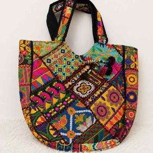 May include: A tote bag featuring a vibrant patchwork design with multi-coloured embroidered patterns, including floral motifs and stylised birds. The bag has black fabric handles and decorative tassels with small beads attached to the front panel.