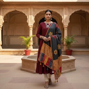 May include: A woman wearing a maroon-colored dress with long sleeves and a patterned shawl. The shawl is primarily black with floral and geometric designs in yellow, red, and blue. The outfit is accessorized with silver jewelry.