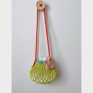 May include: A handmade crochet bag in a light green net design, with a white interior and a pink flower accent. The bag has orange straps and is suspended from a wooden button on a white wall. The bag is approximately 6 inches wide.