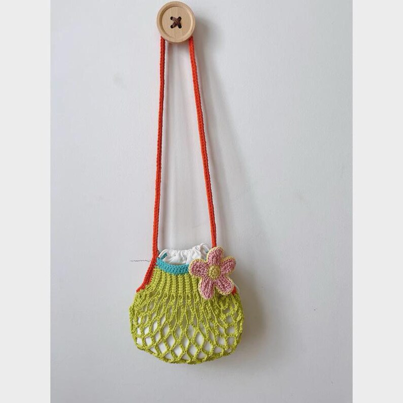 May include: A handmade crochet bag in a light green net design, with a white interior and a pink flower accent. The bag has orange straps and is suspended from a wooden button on a white wall. The bag is approximately 6 inches wide.