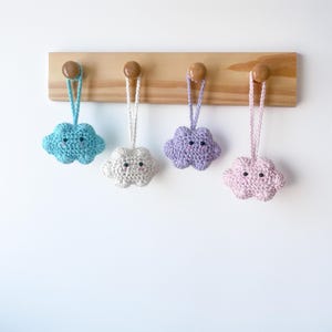 Puede incluir: Cuatro adornos de nube de ganchillo hechos a mano en colores pastel: azul, blanco, morado y rosa. Cada nube tiene una cara sonriente y un lazo para colgar. Se exhiben en un estante de madera con clavijas de madera.
