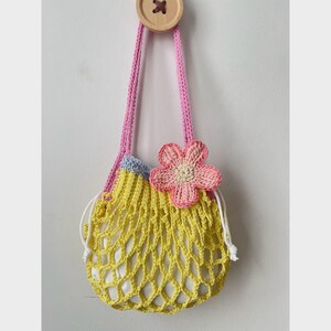 May include: A handmade crochet bag in yellow with a pink strap and a pink flower accent. The bag has a drawstring closure and is hanging from a wooden button. The bag is designed with a net-like pattern.