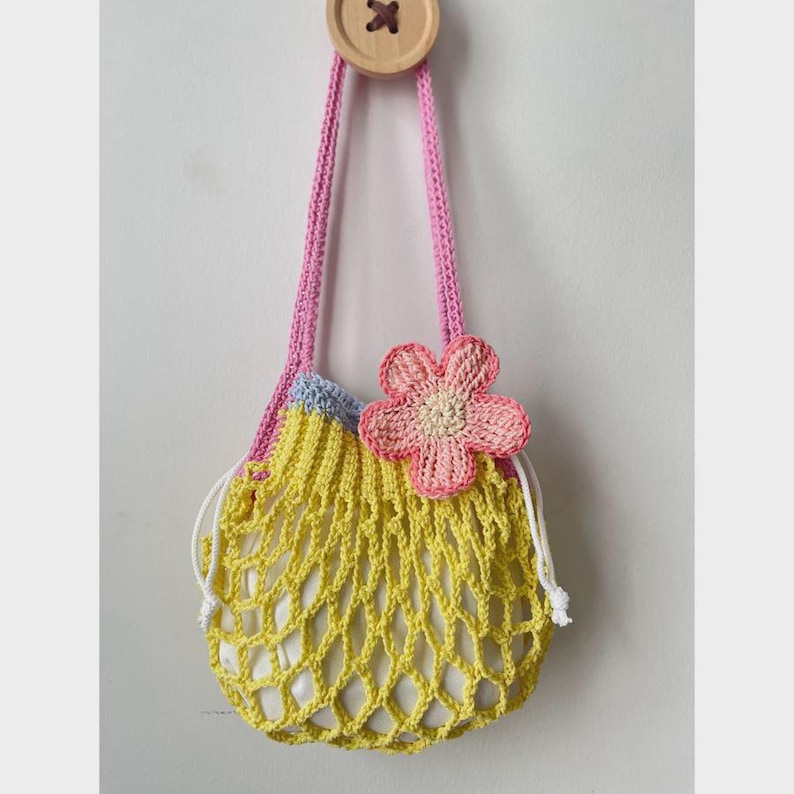 May include: A handmade crochet bag in yellow with a pink strap and a pink flower accent. The bag has a drawstring closure and is hanging from a wooden button. The bag is designed with a net-like pattern.