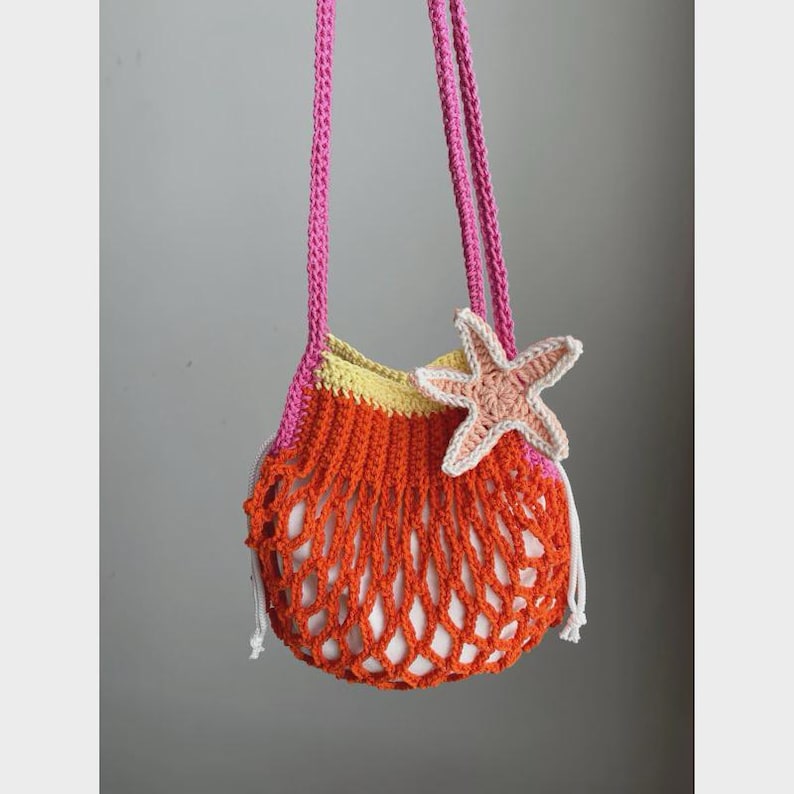 May include: A handmade crochet bag in orange, yellow, and pink, featuring a starfish applique. The bag has a net-like design and long pink straps. The bag is designed to be carried over the shoulder.