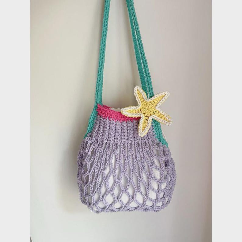 May include: A handmade crochet bag in shades of lavender, teal, and pink, adorned with a yellow starfish. The bag features a net-like design and long teal straps. A whimsical, beach-themed accessory.