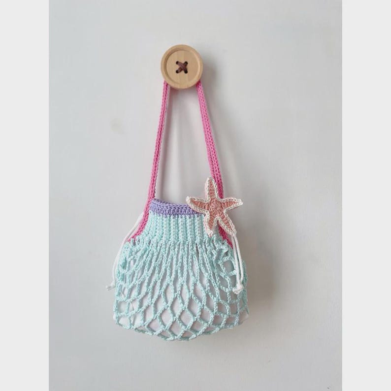 May include: A handmade crochet bag in a light blue net design, with a pink strap and a light purple top. A pink starfish is attached to the bag. The bag is hanging from a wooden button on a white wall. The bag is a beach-themed accessory.