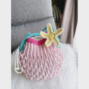 May include: A handmade crochet bag in shades of pink, white, and turquoise, adorned with a yellow and white starfish. The bag features a net-like design and a drawstring closure. The bag is sitting on a white fluffy surface.
