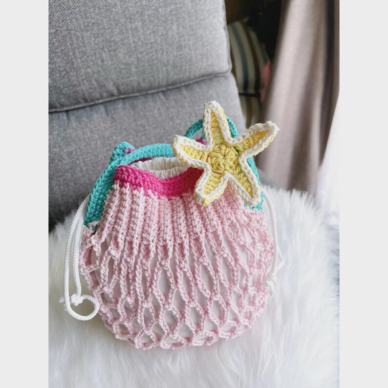 May include: A handmade crochet bag in shades of pink, white, and turquoise, adorned with a yellow and white starfish. The bag features a net-like design and a drawstring closure. The bag is sitting on a white fluffy surface.