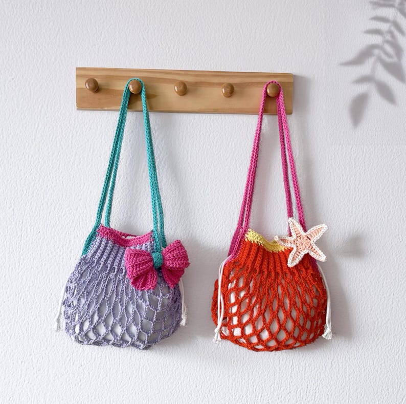 May include: Two handmade crochet net bags hanging on a wooden rack. One is lavender with a pink bow and teal straps, the other is orange with a starfish and pink straps. The bags are designed with a drawstring closure.