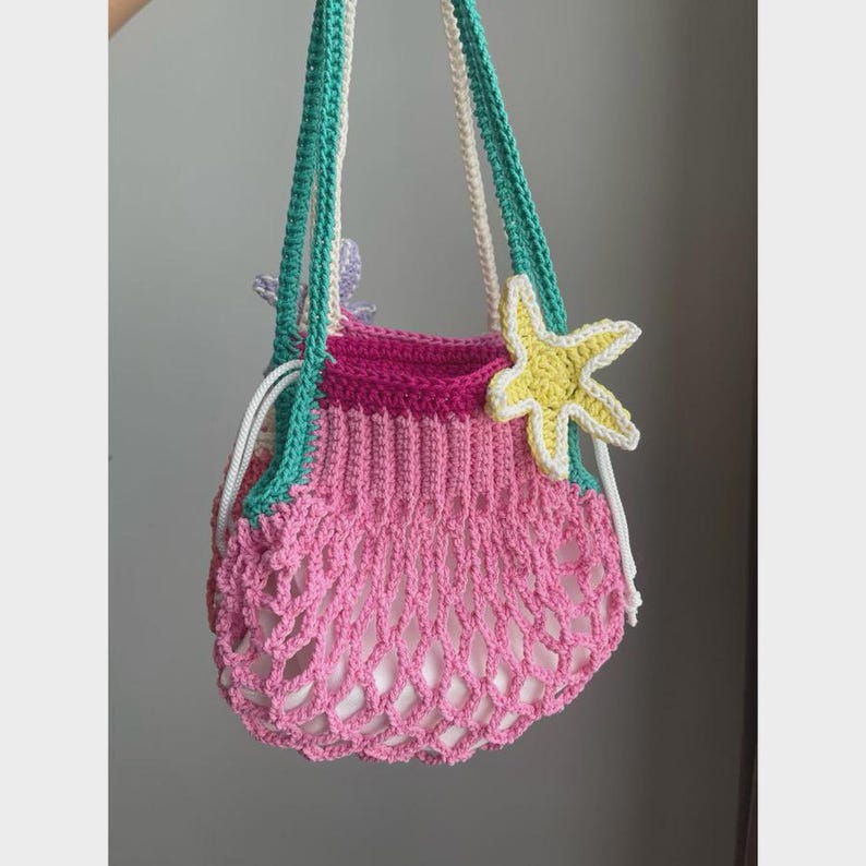 May include: A handmade crochet bag in shades of pink, fuchsia, and teal, adorned with a yellow starfish and a white drawstring. The bag features a net-like design and is held by teal and white straps. A whimsical, beach-themed accessory.