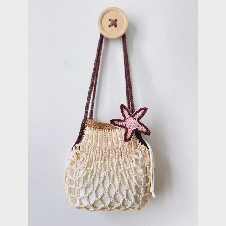 May include: A crocheted net bag in cream and brown, adorned with a pink and brown starfish. The bag hangs from a wooden button attached to the wall by two brown straps.