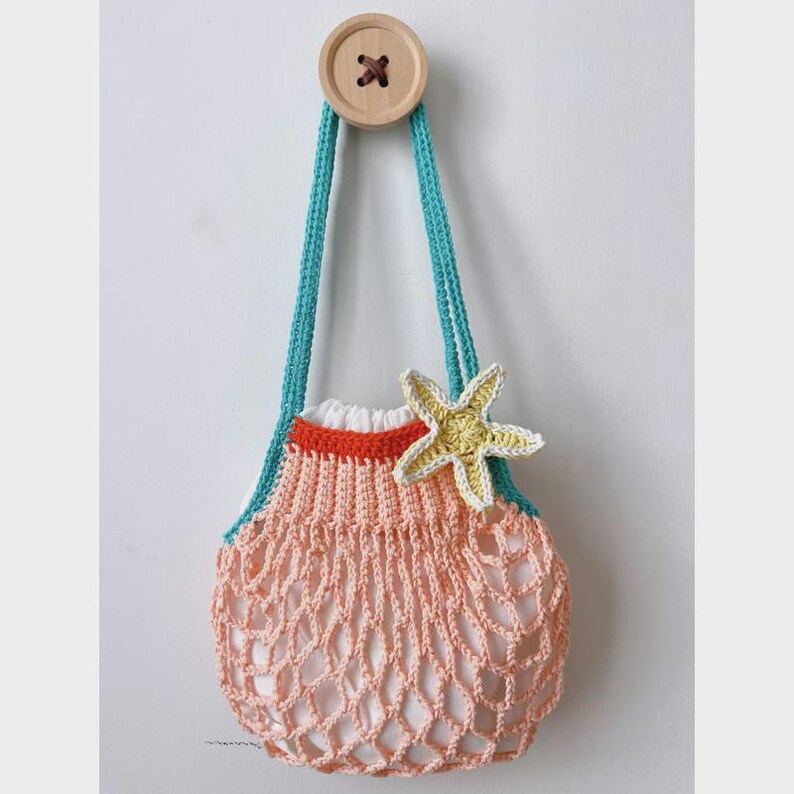 May include: A handmade, crocheted beach bag in a net-like pattern. The bag is peach-colored with turquoise straps and a yellow starfish accent. A wooden button secures the bag to the wall. The bag has a white lining and a red trim.