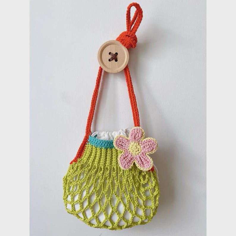 May include: A handmade crochet bag in a light green net design, adorned with a pink and yellow flower. It features orange straps and a wooden button closure. The bag is designed to hang, showcasing a unique, handcrafted aesthetic.