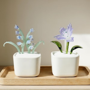 May include: Two decorative glass flower arrangements in white square pots sit on a wooden tray. One features blue bell-shaped flowers, the other a purple flower. The glass stems and leaves are green.
