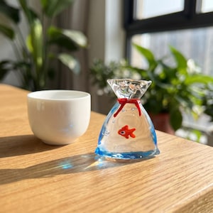 May include: A clear glass goldfish sculpture in a bag, with a red bow. The bag has blue detailing and a small red goldfish inside. A white ceramic cup is next to the sculpture.