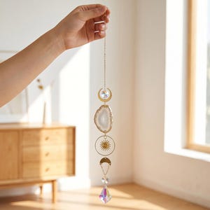 Op de afbeelding: Een hangend zonnevanger-ornament met een goudkleurige metalen ketting, een halvemaanvormige bedel, een doorschijnende witte agaatplak, een ronde zonnebedel, nog een halve maan en een gefacetteerd iriserend kristalprisma aan de onderkant.