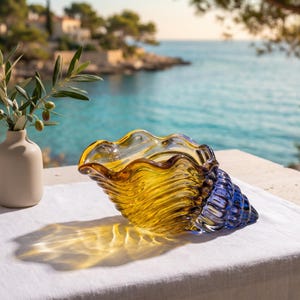Puede incluir: Un cuenco decorativo de cristal con forma de caracola, que presenta un degradado de tonos ámbar y amarillo translúcidos que transicionan a un azul intenso en la base en espiral, con un borde ondulado y un acabado liso y brillante.