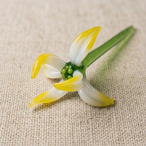May include: A glass flower sculpture with yellow and white petals and a green stem. The centre of the flower has small green spheres. The artwork is set against a neutral background. The petals have yellow tips.