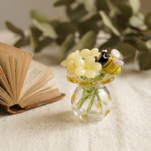Puede incluir: Un jarrón miniatura de vidrio transparente que contiene un pequeño ramo de flores de vidrio amarillo pálido y una figura de abeja de vidrio con rayas amarillas y negras, alas transparentes y cabeza negra, todo con superficies lisas.