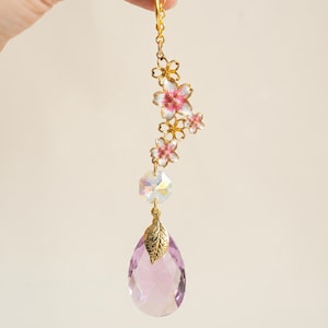 Könnte beinhalten: Ein dekorativer Hängeanhänger mit einer goldfarbenen Kette, einer Ansammlung von weiß-rosa Emaille-Kirschblüten, einer klaren facettierten Kristallperle und einem großen, tropfenförmigen hellvioletten Kristallanhänger mit einem goldenen Blatt.