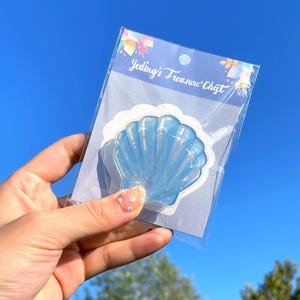 Op de afbeelding: Een blauwe schelpvormige sticker met glitteraccenten, verpakt in een transparante plastic hoes. De tekst "Yeding's Treasure Chest" staat op de verpakking. De sticker wordt tegen een helderblauwe lucht gehouden.