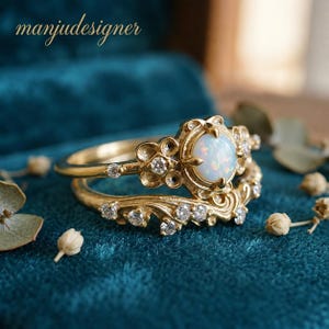 May include: A gold ring set featuring a central round opal cabochon surrounded by small clear stones and intricate metalwork. The band includes a curved, decorative piece adorned with additional clear stones. The text manjudesigner is visible.