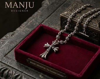 May include: A silver cross pendant necklace with a detailed design, displayed in a red velvet jewelry box. The necklace is part of "The Guardian Collection" by Manju Designer, as indicated by the text on the box.