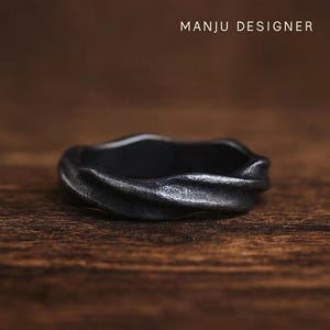 May include: A dark grey ring with a twisted design, set against a wooden background. The ring has a textured surface. The text "MANJU DESIGNER" is visible at the top.