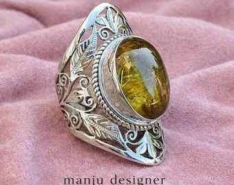May include: A silver-toned ring with an oval, yellow-green gemstone. The ring features intricate leaf and swirl designs on the band and around the gemstone. The text "manju designer" is at the bottom of the image.