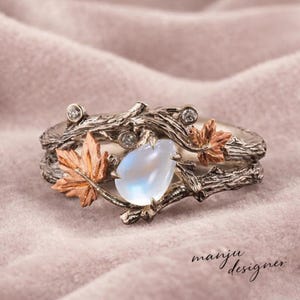 May include: A silver ring with a nature-inspired design. The ring features a pear-shaped, light blue gemstone, accented with small diamonds and copper-colored maple leaves. The band is crafted to resemble tree branches. The text "manju designer" is visible.