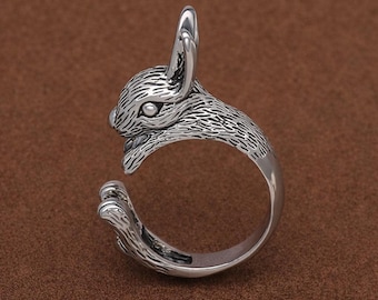 May include: A silver-colored ring in the shape of a rabbit. The rabbit's head and one ear are upright, with the ring band forming the rest of the body. The ring has detailed textures and is set against a brown background. The text "manju designer" is at the bottom.