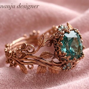 May include: A rose gold ring with an elaborate leaf design and a large, square, teal gemstone. The ring has the text "manju designer". The ring is set on a pink surface.