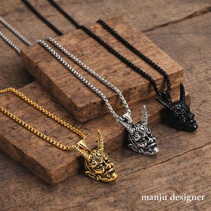 May include: Four necklaces with pendant designs. The pendants are shaped like a horned mask. One necklace is gold, one is silver, and one is black. The necklaces are on a wooden surface. The text "manju designer" is visible.