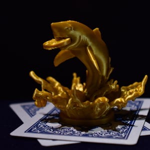 Puede incluir: Una escultura dorada de un tiburón saltando fuera del agua, sosteniendo una moneda en la boca. La escultura está colocada sobre dos cartas de juego con dorsos estampados en azul y blanco. El fondo es oscuro.