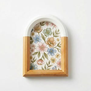 Pode incluir: Uma bandeja decorativa de madeira em forma de arco com parte superior branca e laterais de madeira natural. A bandeja apresenta um padrão floral com flores rosa, azuis e bege em fundo branco. A bandeja é decorativa.