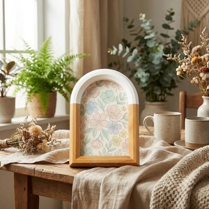 May include: A wooden arch-shaped picture frame with a white top and natural wood sides. The frame holds a floral print with pastel pink, blue, and yellow flowers. The frame is displayed on a wooden table with other decorative items.