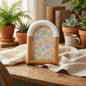 May include: A wooden picture frame with an arched top, painted white, and a natural wood base. The frame holds a floral print with pastel pink, blue, and yellow flowers. The frame is displayed on a wooden table with plants in terracotta pots.